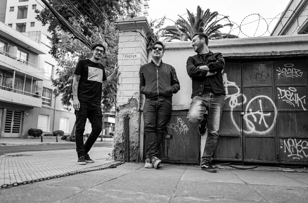 Los tres integrantes de la banda apoyados y conversando junto a un muro de la calle, con una cadena en el piso