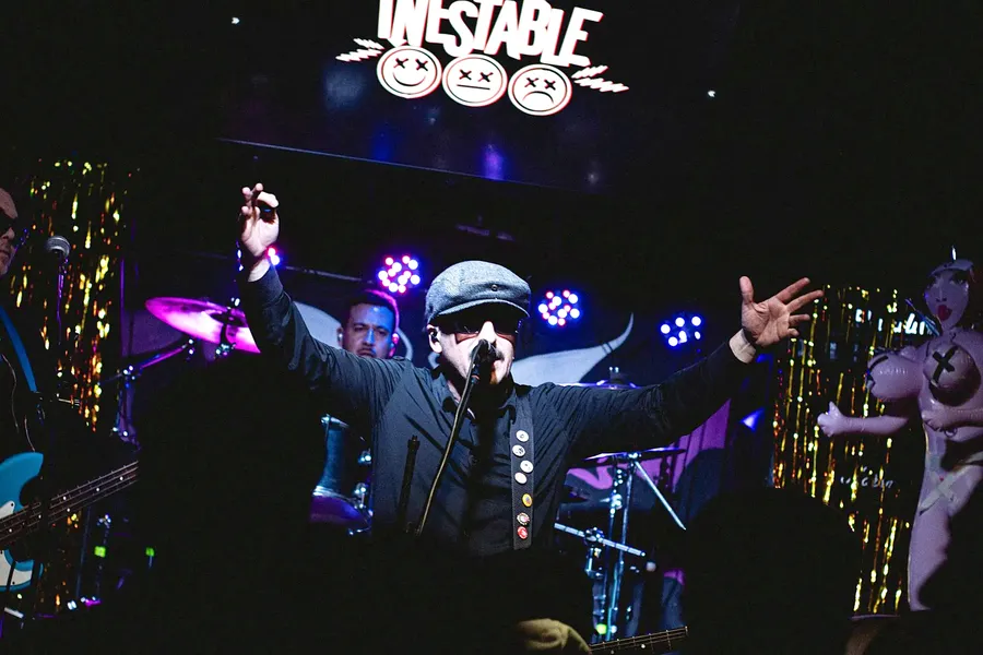 Cantante con gorra y lentes oscuros, con los brazos abiertos en el escenario, bajo el logo de Inestable